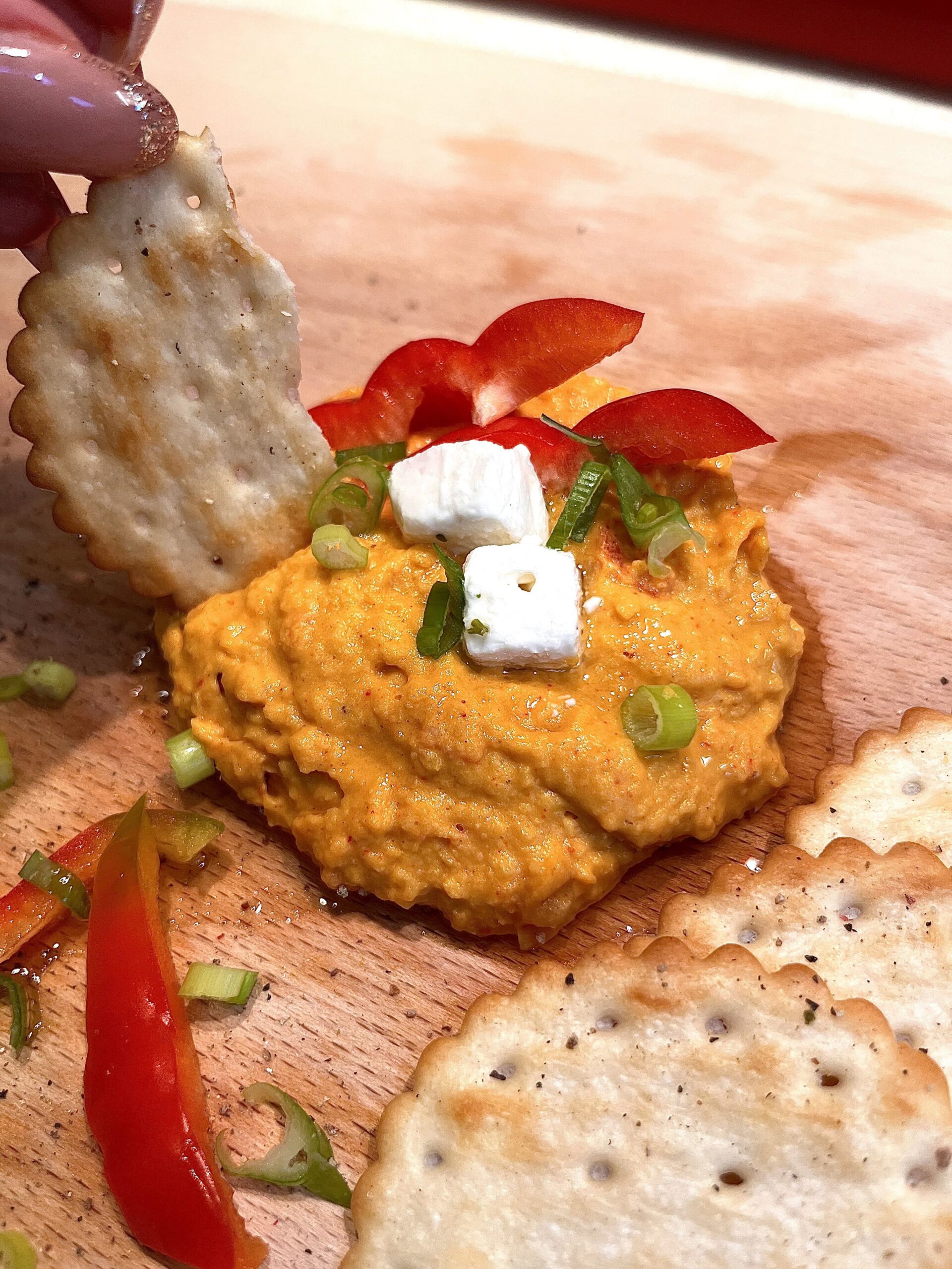 Homemade easy hummus recipe served on a wooden board, topped with feta cubes, red bell pepper slices, spring onions, and olive oil, accompanied by crispy crackers. Perfect for a quick and easy healthy snack.