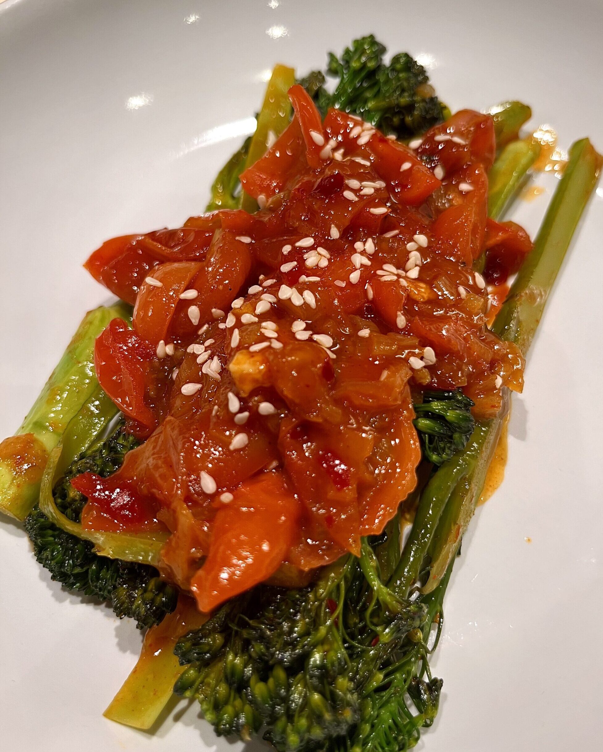 Sautéed tenderstem broccoli with sweet chilli, featuring vibrant green broccoli, cherry tomatoes, shallots, and garlic, coated in a glossy sweet chilli glaze and garnished with sesame seeds, served as a quick and easy side dish