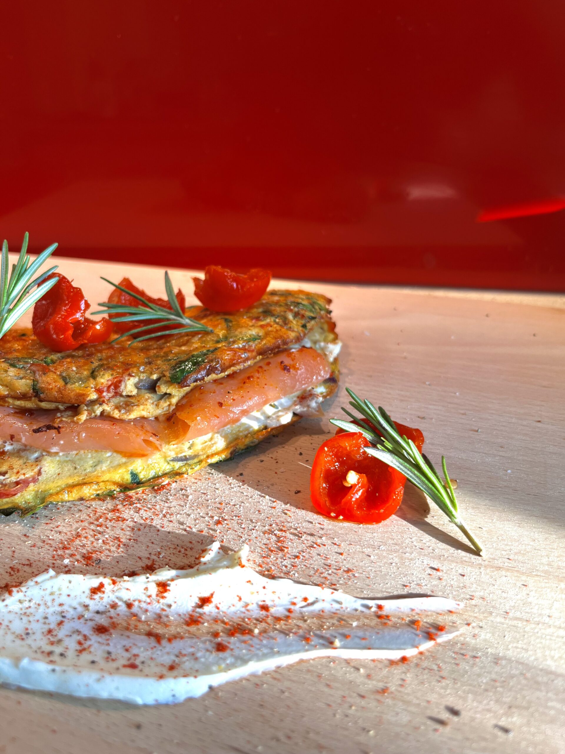 Fluffy, golden omelette filled with creamy cream cheese and rich smoked salmon, garnished with fresh rosemary and vibrant cherry peppers, served on a wooden board. A high-protein, indulgent breakfast or brunch dish.