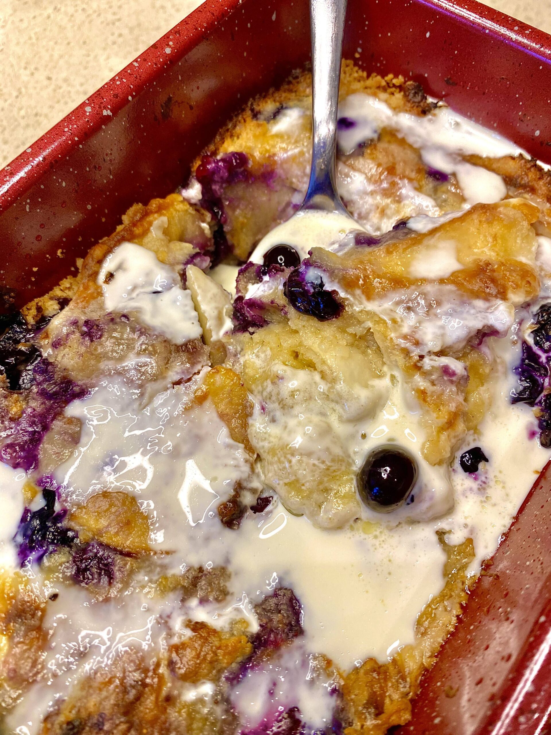 Homemade blueberry peach cobbler with a golden crust, topped with cream drizzled on top, served warm for a comforting dessert.